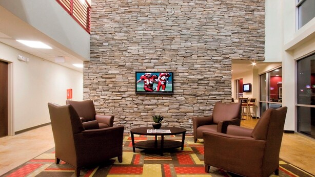 A lobby with a stone wall, two brown armchairs, a small table with a plant, and a television screen. - Red Roof Inn & Suites Beaumont (Beaumont)