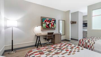 Habitación de hotel con escritorio, dos camas, televisor, lámpara y vista a la cocina.
