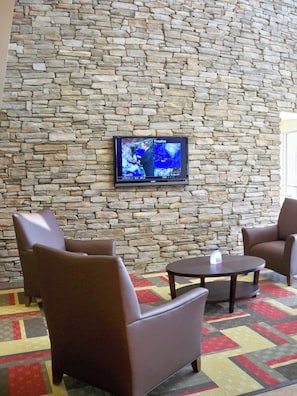A stone wall with a flat-screen TV displaying a weather forecast. There are two brown armchairs and a small round table with a remote control on it. - Red Roof Inn & Suites Beaumont (Beaumont)