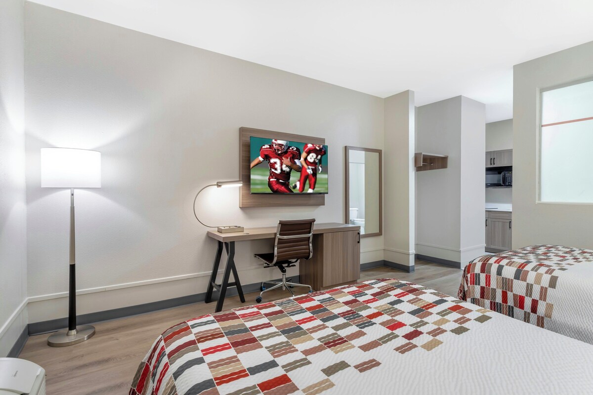 A hotel room with a desk, two beds, a TV, a lamp, and a view of the kitchen.