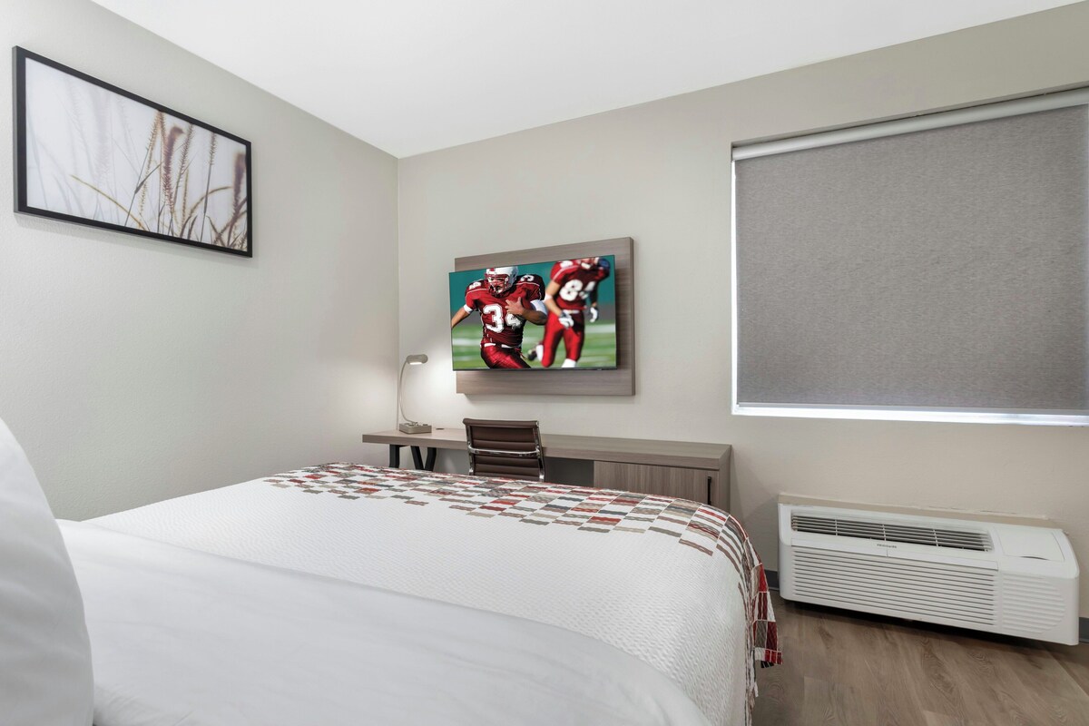 A hotel room with a bed, a desk, a chair, a television, and an air conditioning unit.