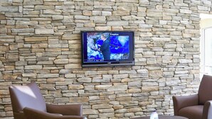 A stone wall with a flat-screen TV displaying a weather forecast. There are two brown armchairs and a small round table with a remote control on it. - Red Roof Inn & Suites Beaumont (Beaumont)