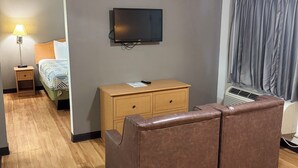 Ein Hotelzimmer mit braunem Ledersofa, einem Nachttisch aus Holz mit Lampe, einem Flachbildfernseher an der Wand, einer Holzkommode und einem Fenster mit Vorhängen.
