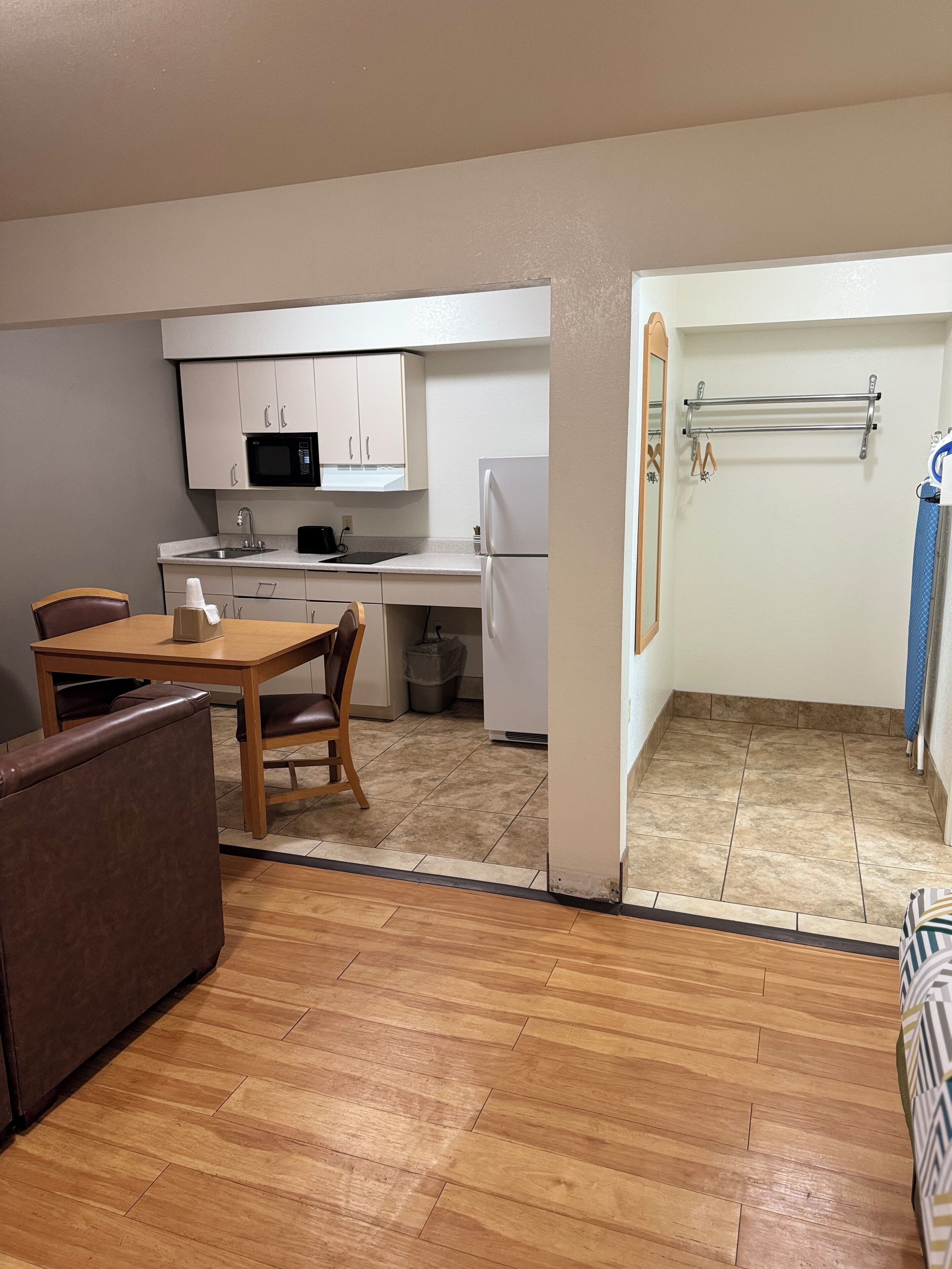 A compact kitchen with white cabinets, a microwave, and a refrigerator. There is a wooden dining table with chairs and a brown sofa. A trash can is visible near the kitchen.