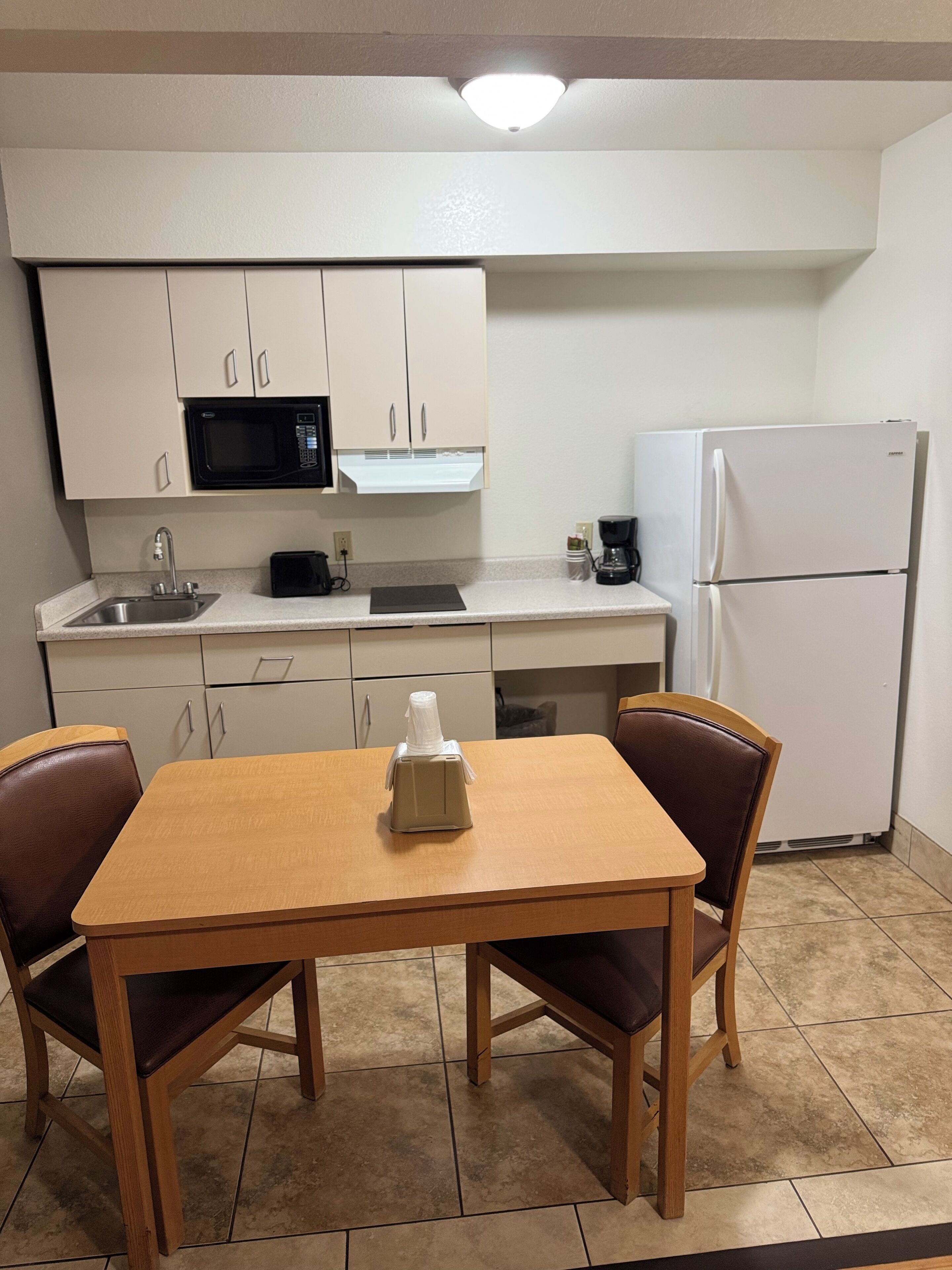 A compact kitchen with white cabinets, a microwave, a coffee maker, and a refrigerator. There is a wooden table with two chairs.