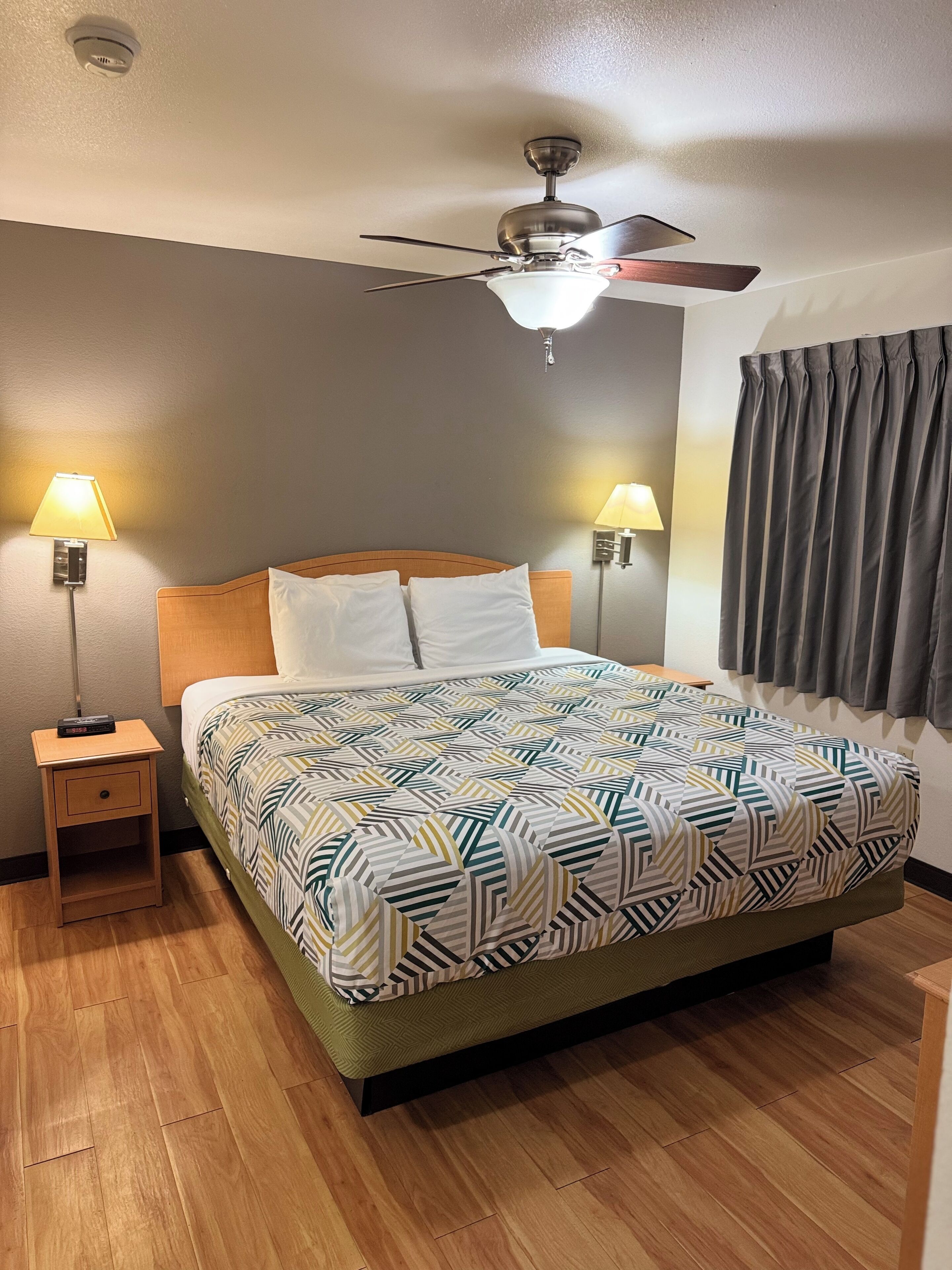A hotel room with a bed, two bedside lamps, a ceiling fan, and a window with curtains.