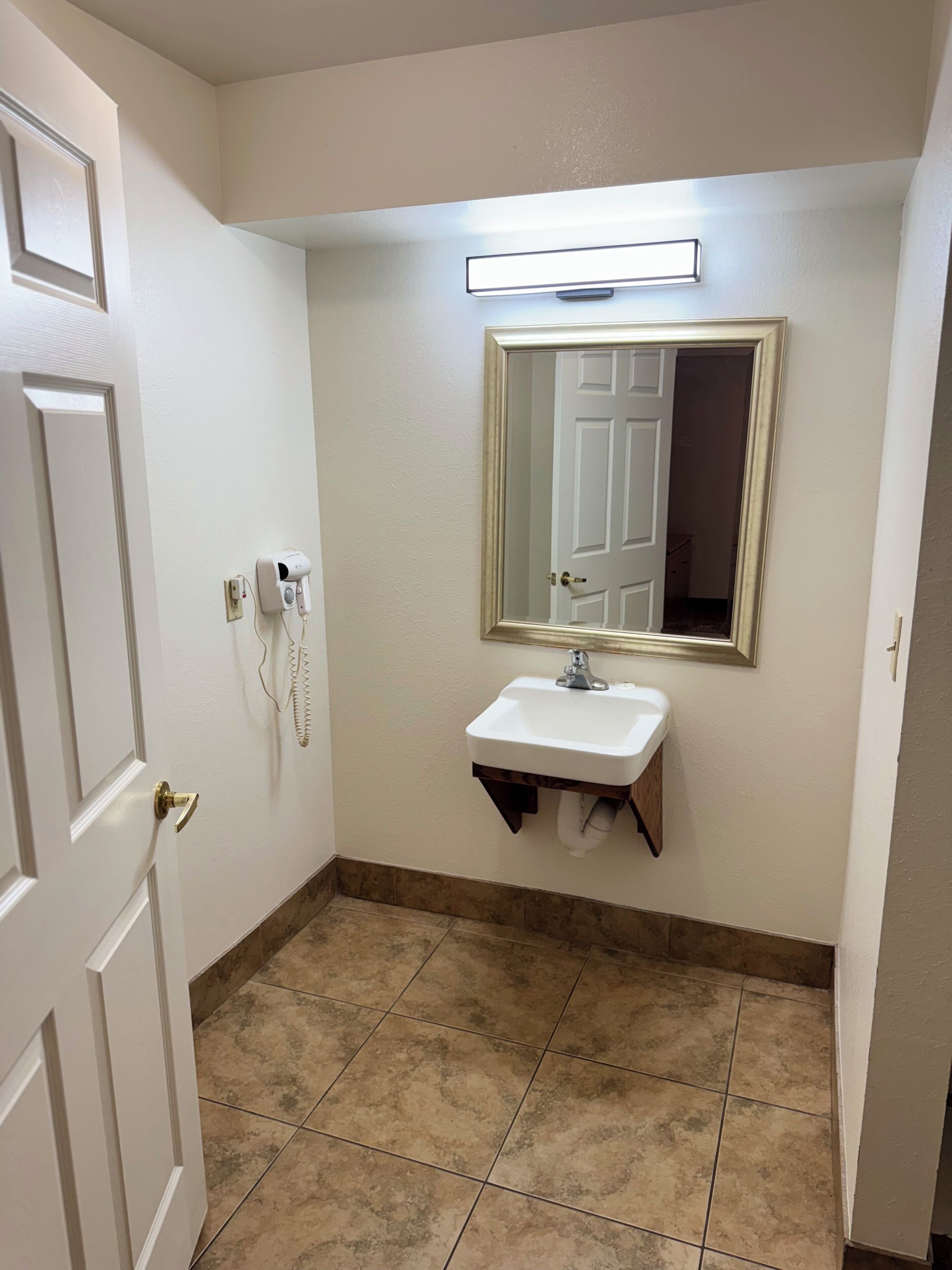 A bathroom with a white door, a white sink with a mirror above it, and a light fixture on the wall.