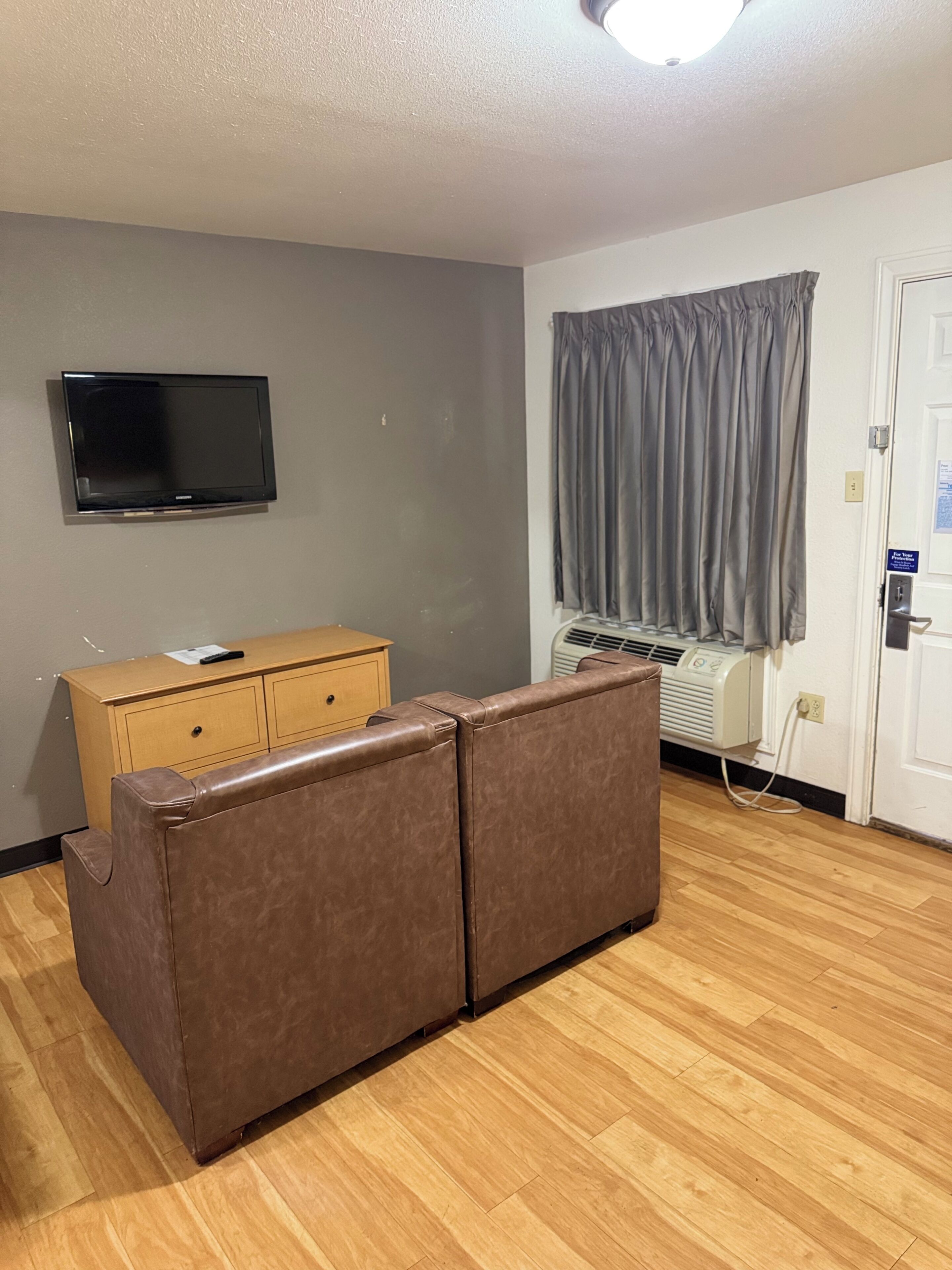 Ein Zimmer mit braunem Ledersofa, einem hölzernen Schminktisch, einem wandmontierten Flachbildfernseher, einem Fenster mit grauen Vorhängen und einer weißen Tür.