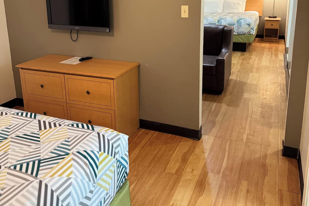 A hotel room with a bed, a wooden dresser, a flat-screen TV, and a hallway leading to another room with a bed and a nightstand.