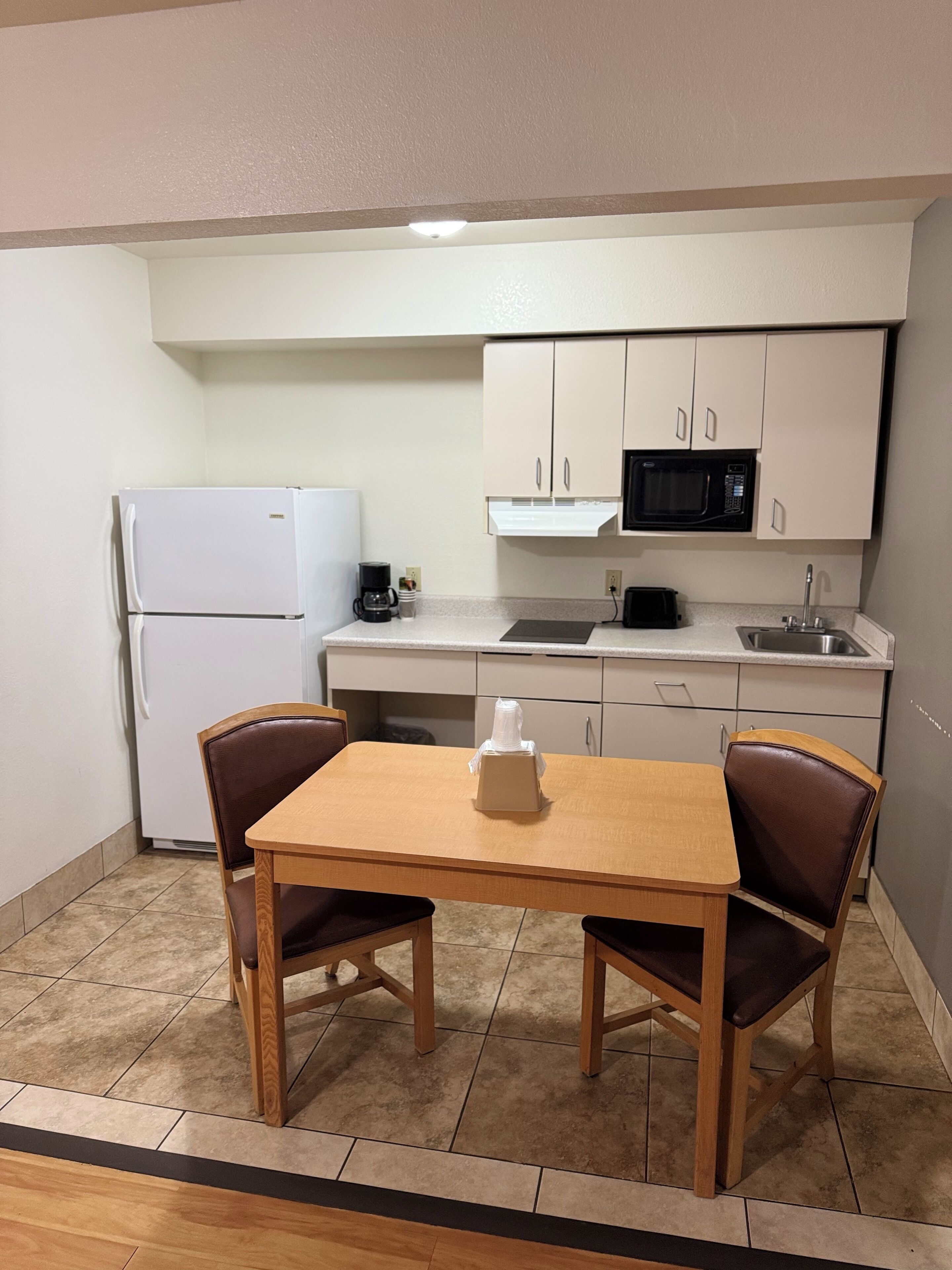 A compact kitchen with a refrigerator, microwave, and sink. There is a wooden table with two chairs and a small white object on it.