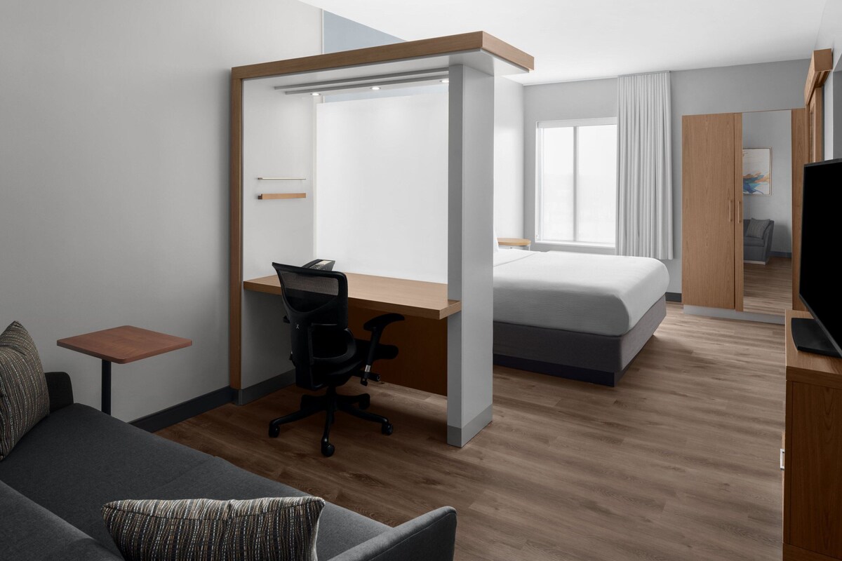 A modern hotel room with a separate desk area, a bed, a sofa, and a television.