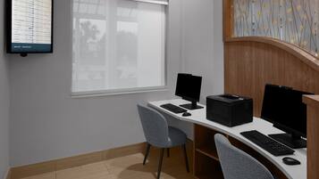 A modern office space with two desks, each equipped with a computer monitor, keyboard, and mouse. There is a large window providing natural light, and a wall-mounted screen displaying information.