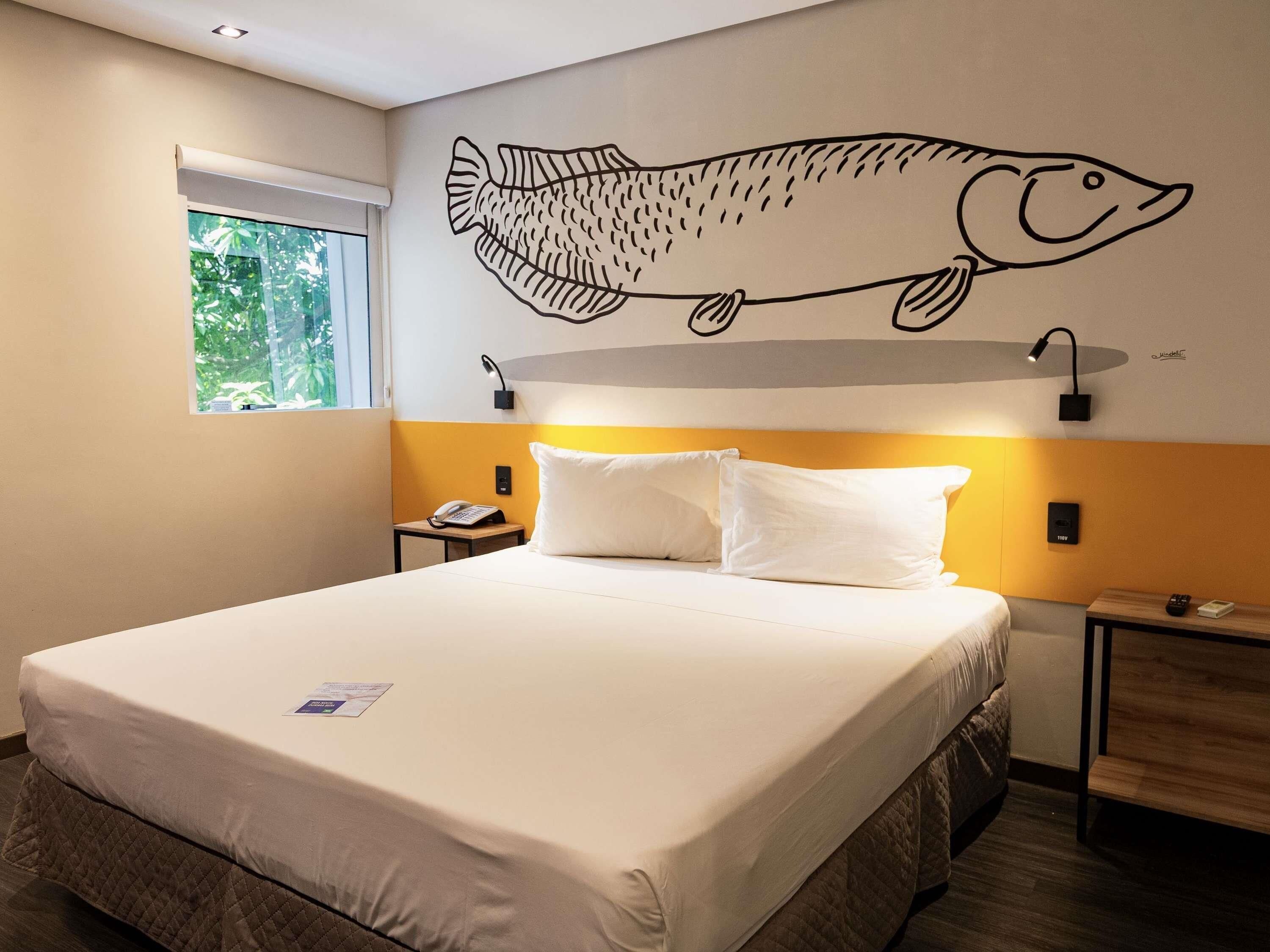 Quarto de hotel com uma cama, um mural de peixe na parede, luminárias de cabeceira e uma janela com vista para vegetação.