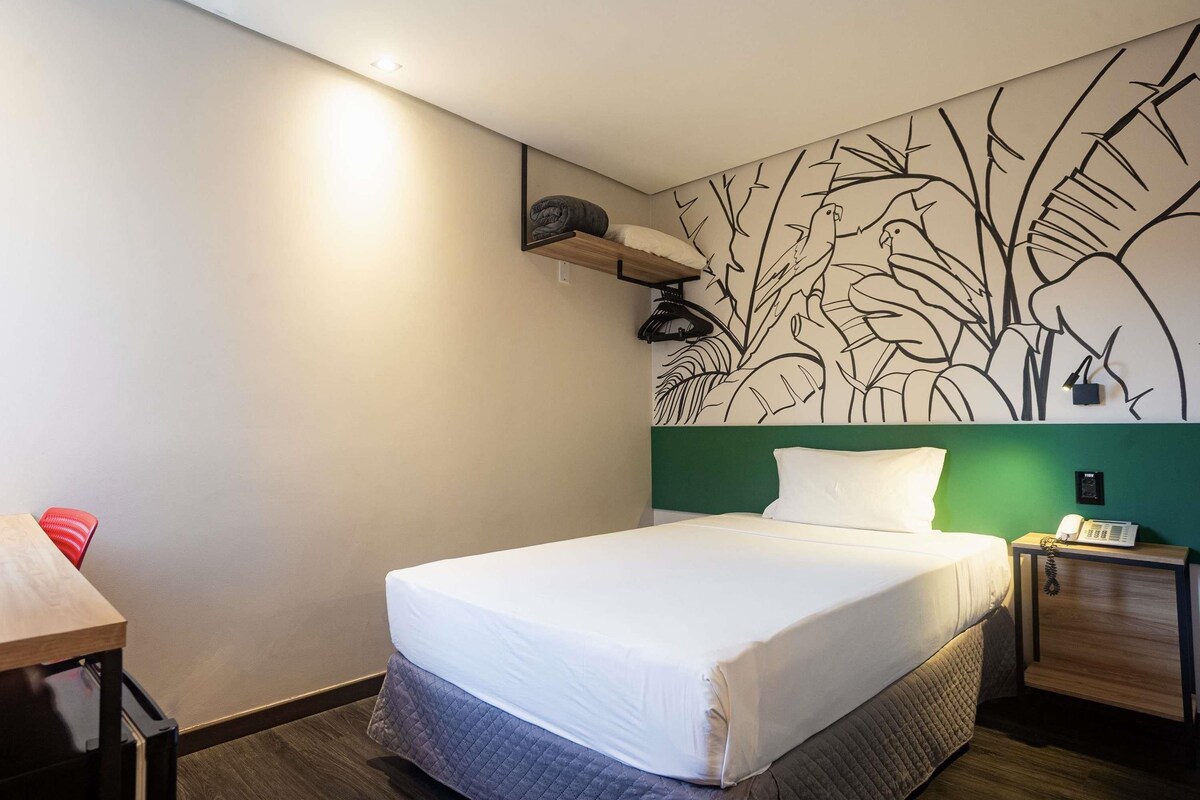Hotellihuone, jossa on sänky, yöpöytä, pieni sivupöytä ja seinä, joka on koristeltu kasvitaihuisella seinämaalauksella.