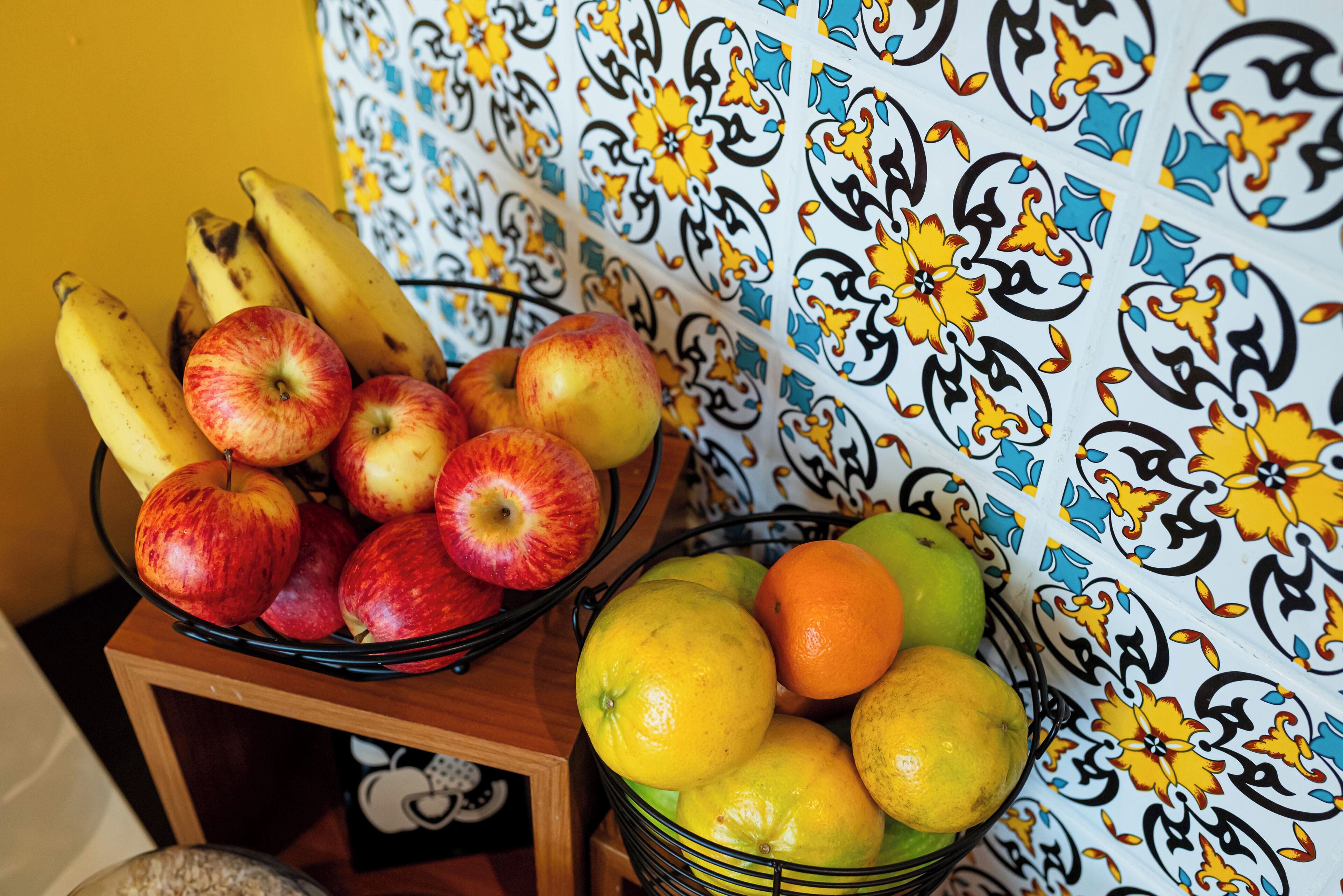 Un frutero con manzanas y plátanos sobre una mesa frente a una pared estampada.