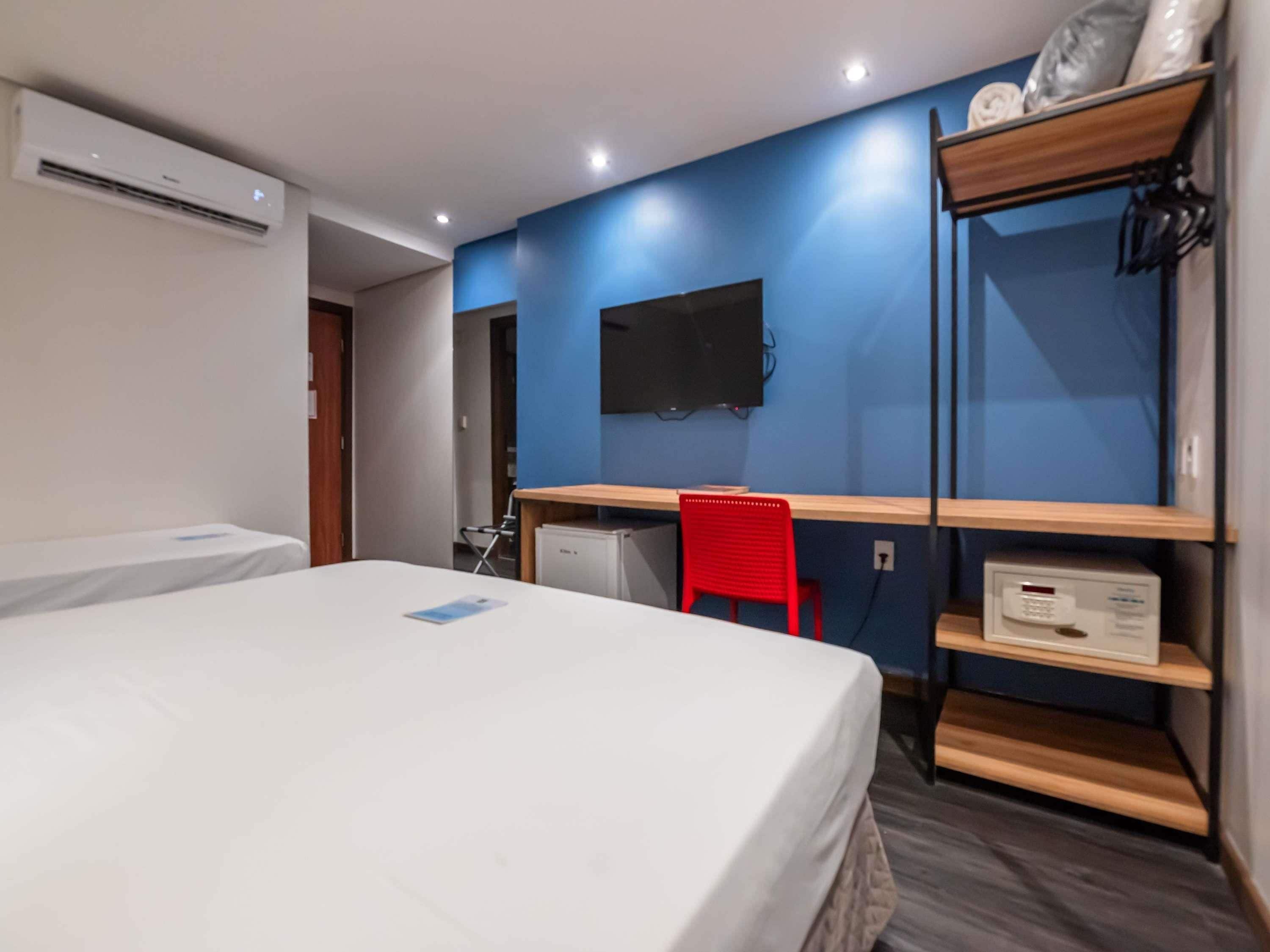 Quarto com cama, escrivaninha, cadeira, televisão e prateleira com objetos.