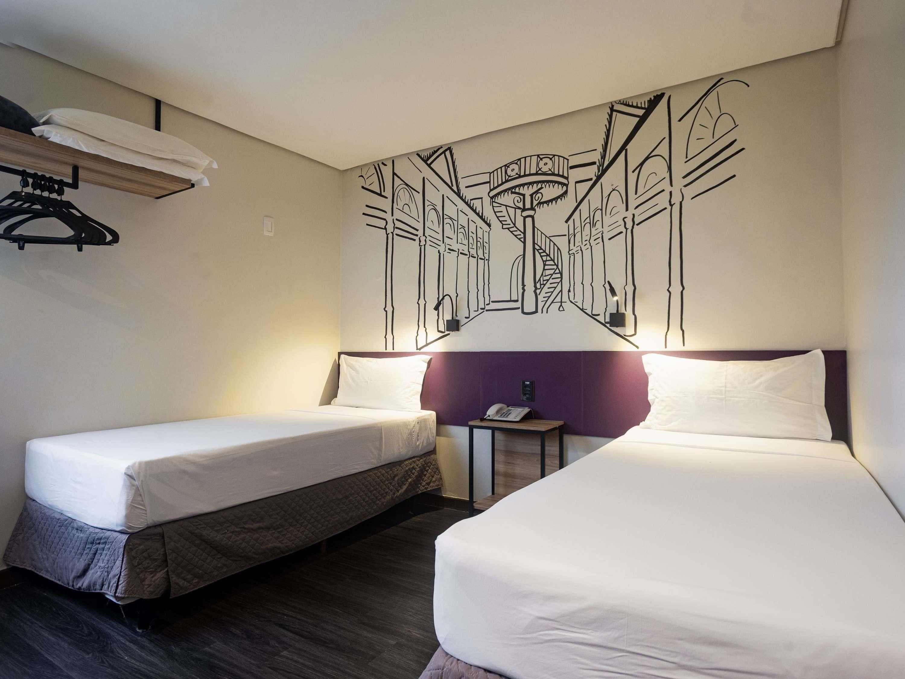 Una habitación de hotel con dos camas individuales, una mesita de noche pequeña con un teléfono, una lámpara de pared y una lámpara en el techo.