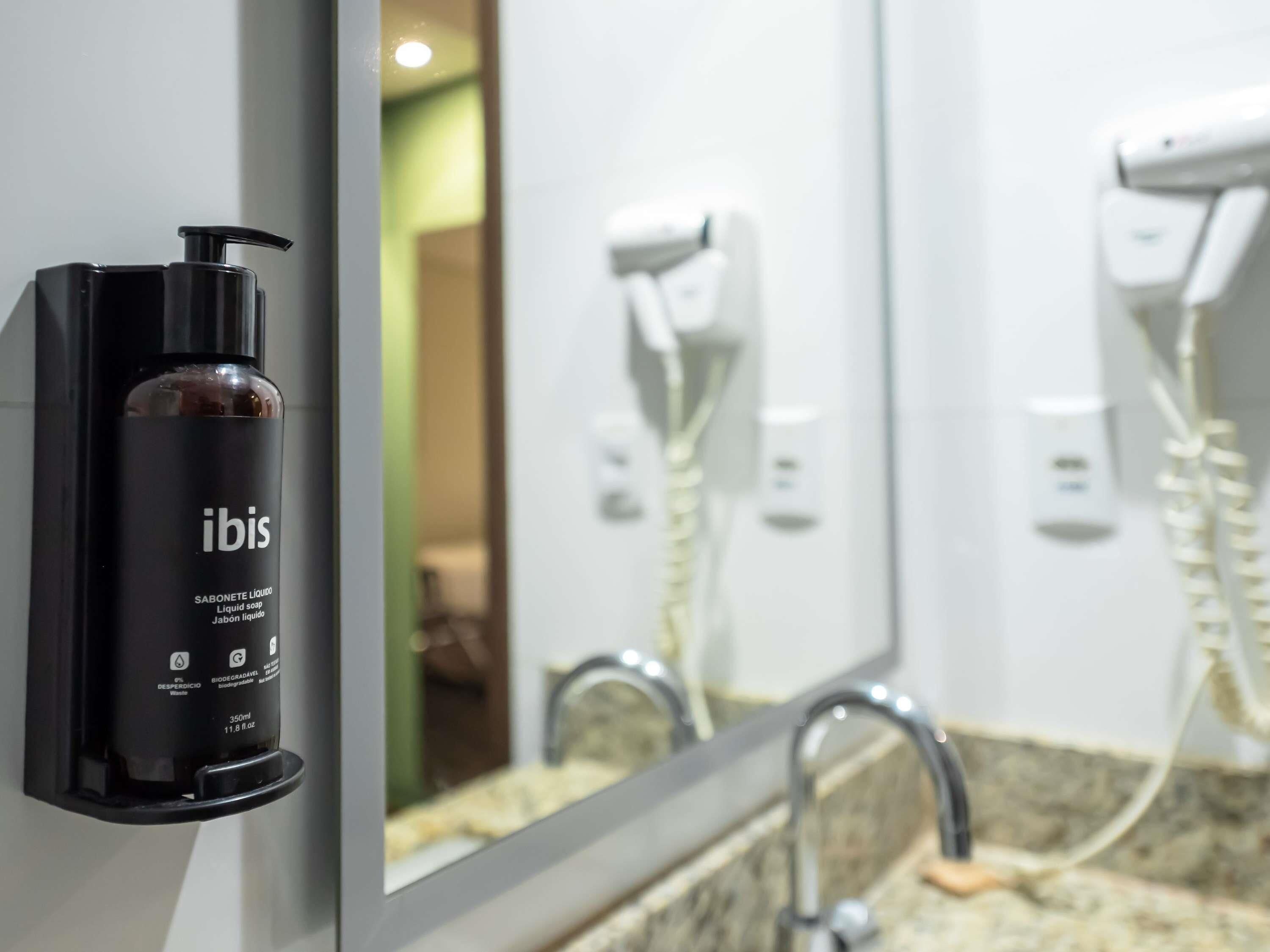 A bathroom with a black bottle of Ibis liquid soap, a mirror, a sink, and a hairdryer.