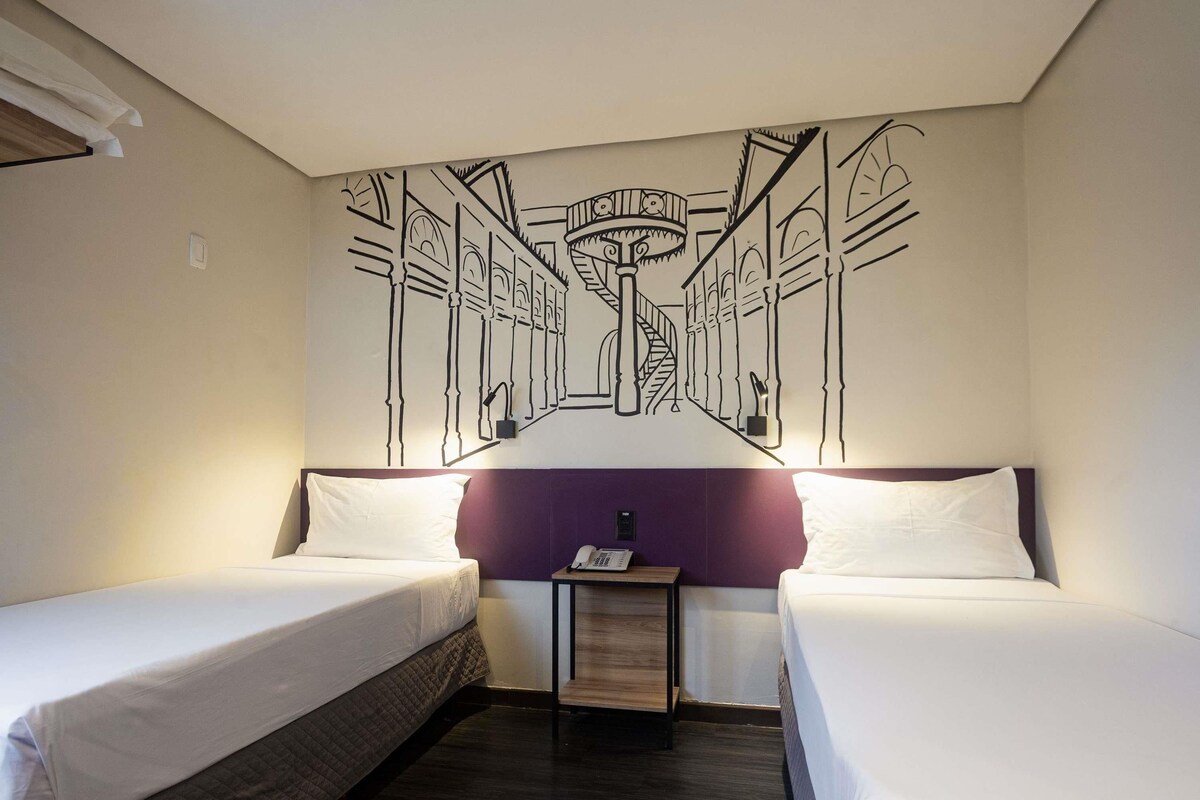 Hotellihuone, jossa on kaksi erillistä sänkyä, pieni yöpöytä lampun kanssa ja seinämaalaus, jossa on kuvattuna tyylitelty rakennus.