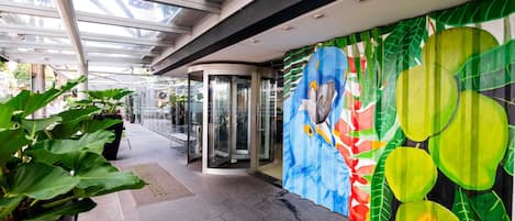 Entrada de hotel com porta de vidro, plantas em vasos e um mural colorido na parede.
