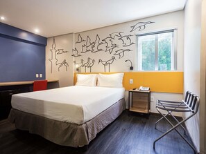 A modern hotel room with a large bed, a desk, a chair, and a window with a view of greenery.