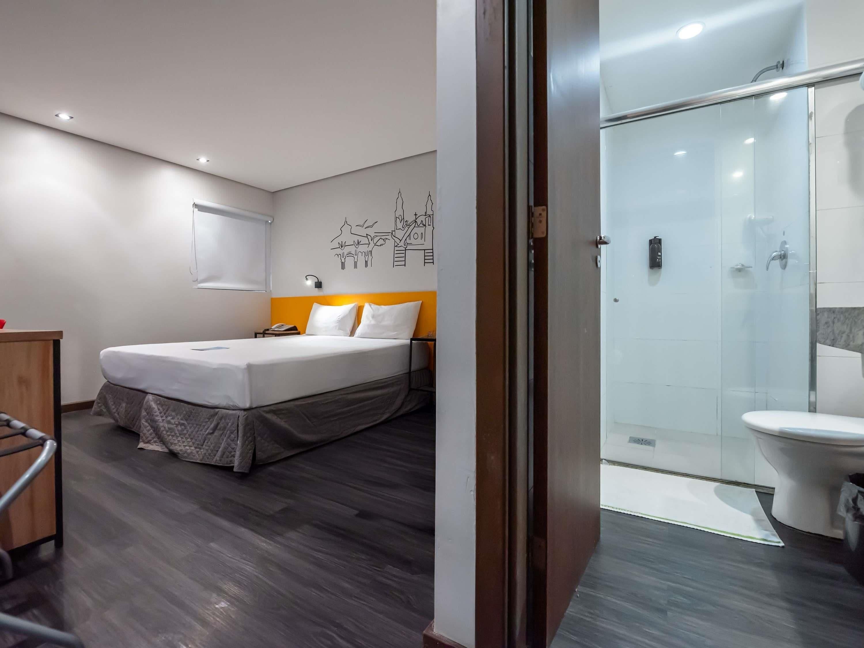 A modern hotel room with a bed, a desk, a shower area, and a toilet.