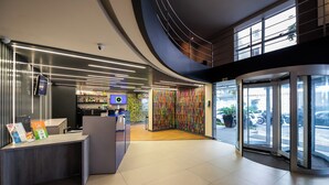 Een moderne hotellobby met een receptiebalie, een digitaal scherm, een kleurrijke muur en een glazen ingang.