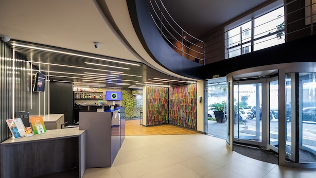 Een moderne hotellobby met een receptiebalie, een digitaal scherm, een kleurrijke muur en een glazen ingang.