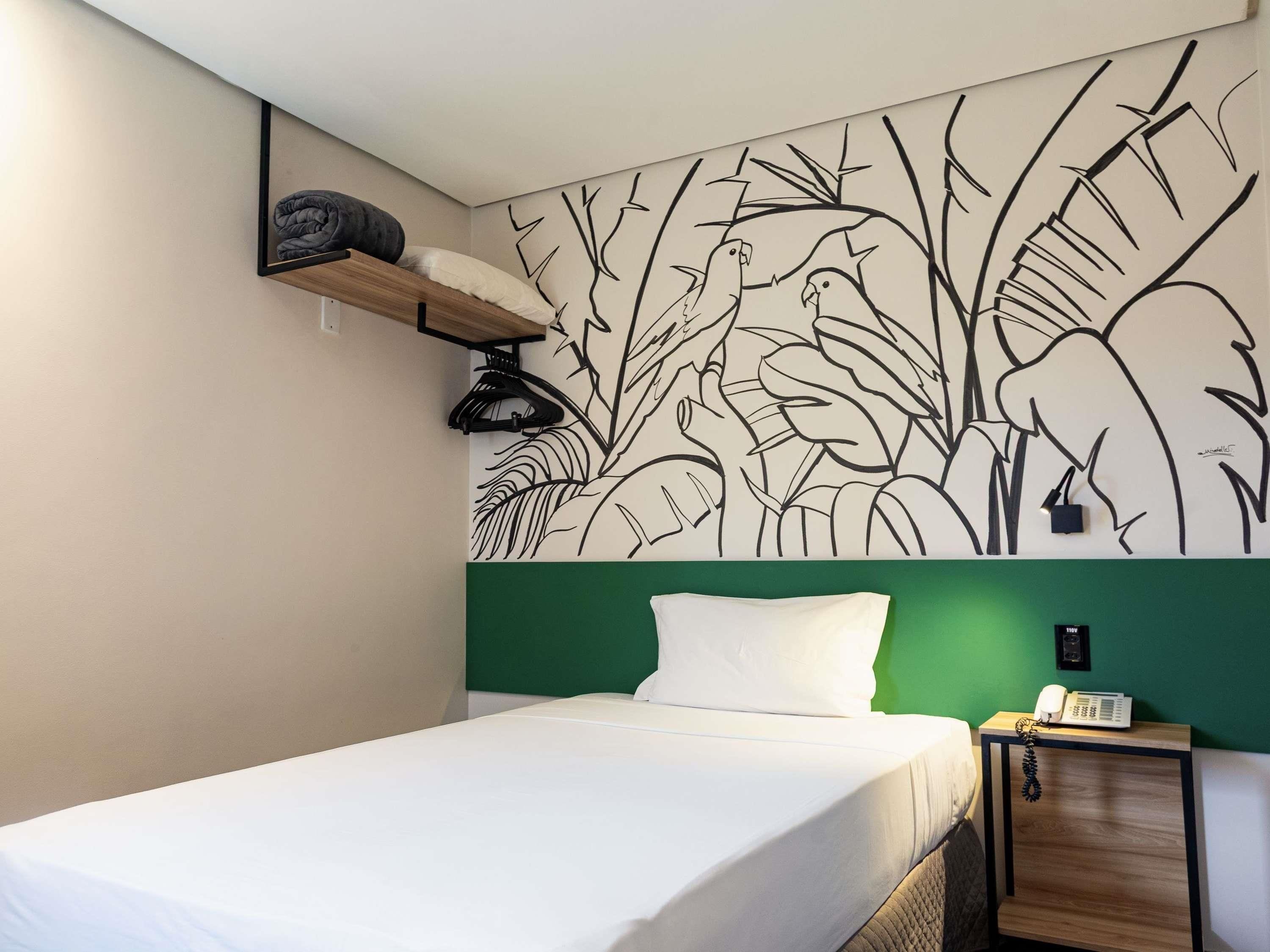 A hotel room with a bed, a green headboard, a wall mural, a bedside table with a phone, and a hanging coat rack.
