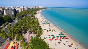 Ein Strand mit Reihn von Sonnenschirmen und Palmen, ein klarer Himmel und Gebäude im Hintergrund.