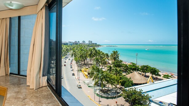 Ein Hotelzimmer mit großem Fenster mit Blick auf einen Strand, Palmen, eine Straße und eine Stadtlandschaft in der Ferne.