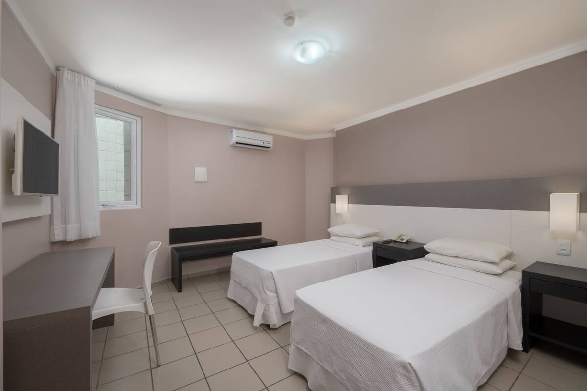 A hotel room with two beds, a desk, a chair, a TV, and an air conditioning unit.