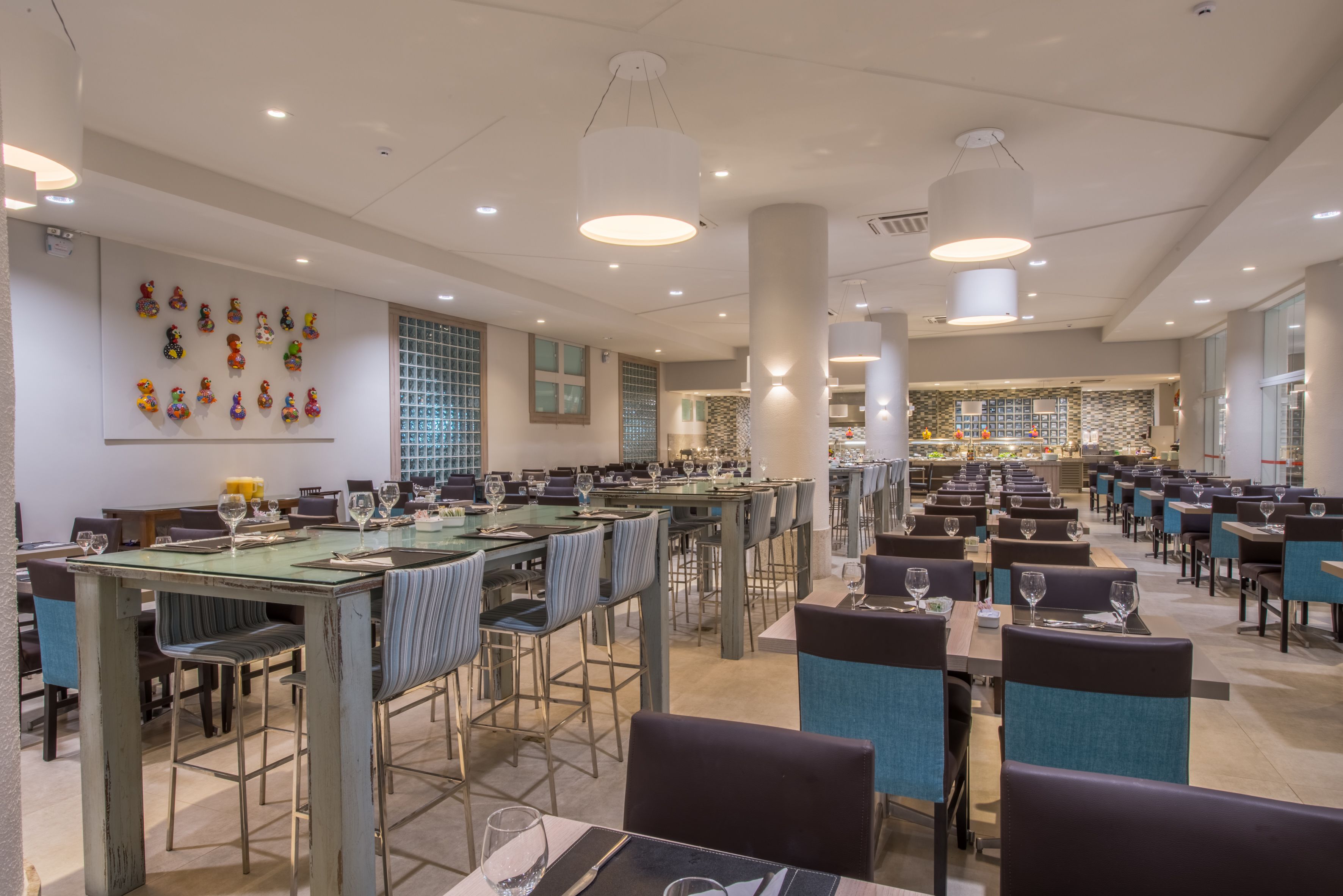 A spacious restaurant with multiple dining tables and chairs, a modern ceiling design, and a decorative wall with colorful items.