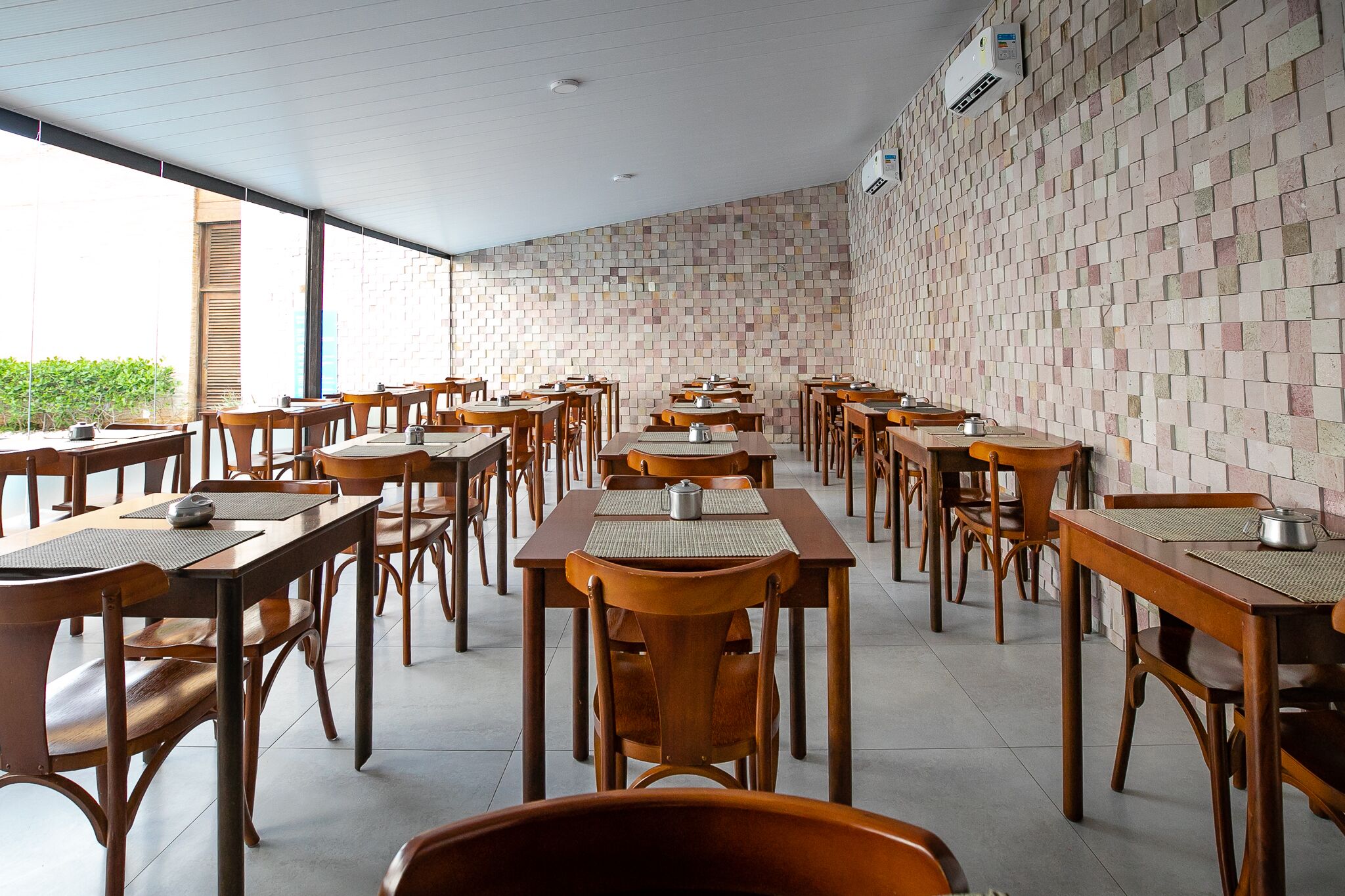 A spacious dining area with wooden tables and chairs, a patterned brick wall, and large windows allowing natural light.