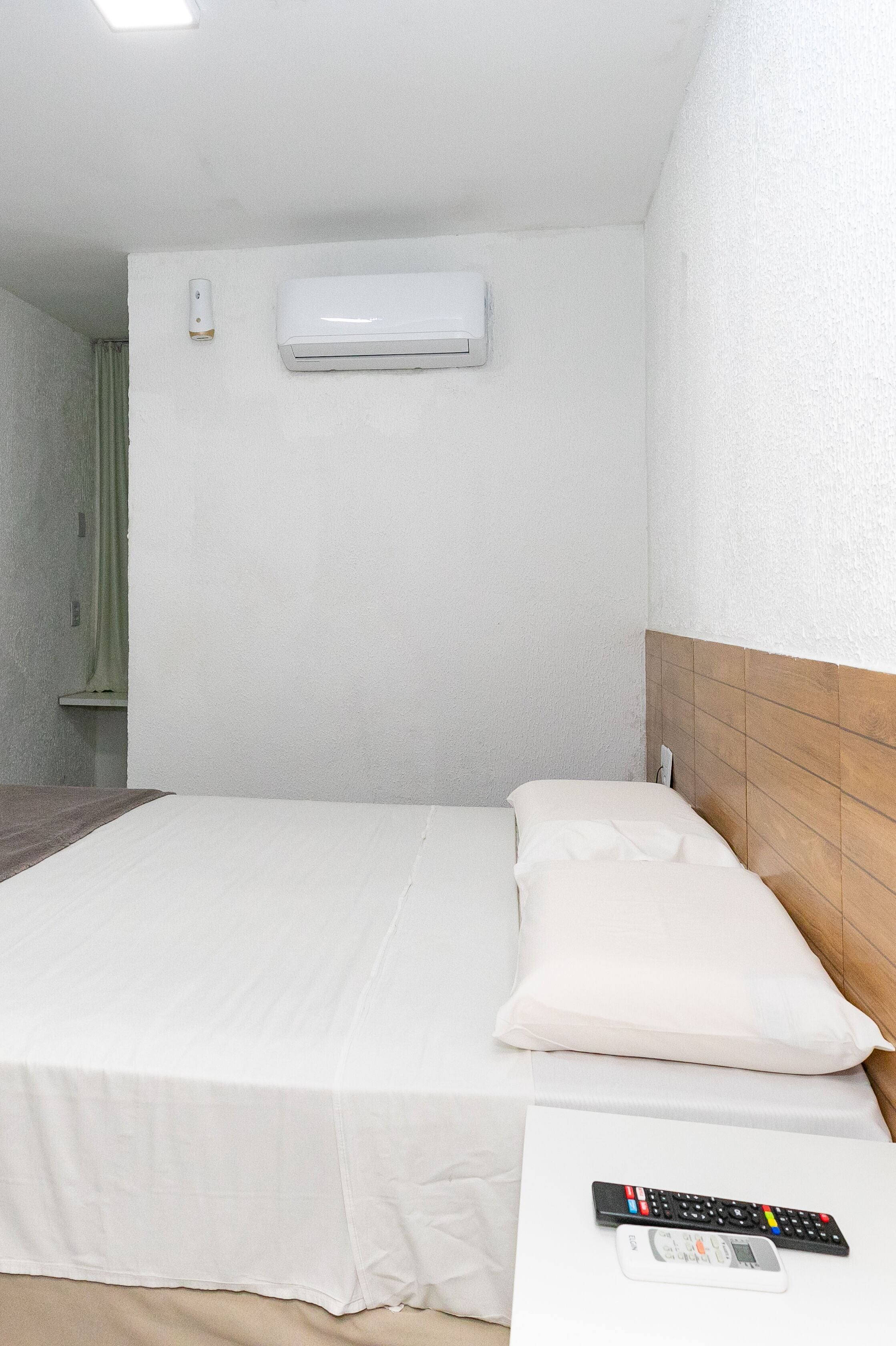 A hotel room with a single bed, a bedside table, a remote control, and an air conditioning unit on the wall.