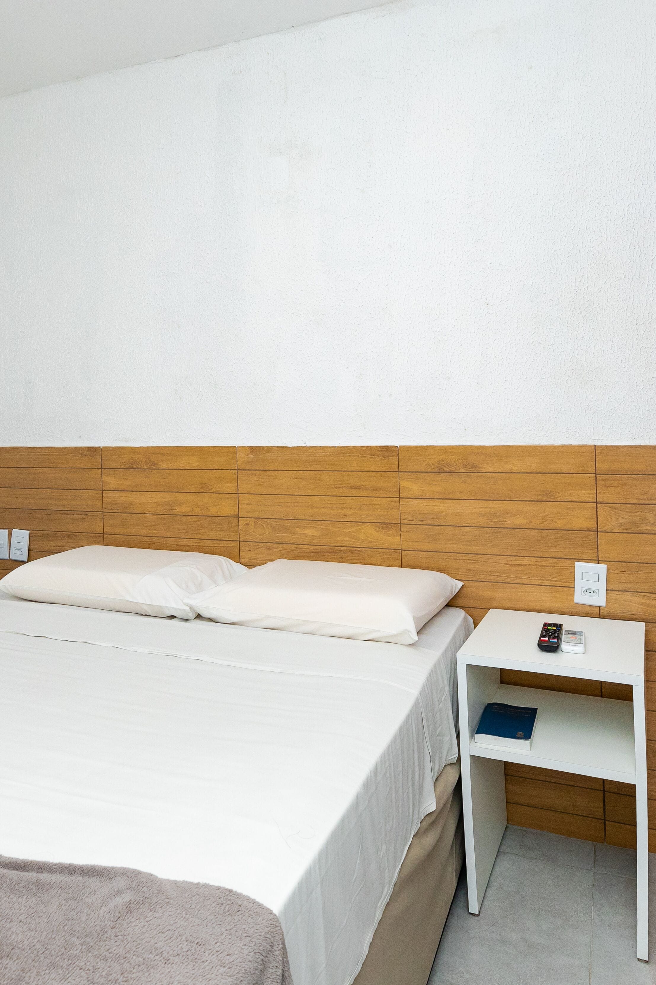 Una cama bien hecha con sábanas blancas, un cabecero de madera, una mesita de noche blanca con un control remoto y un libro azul encima.