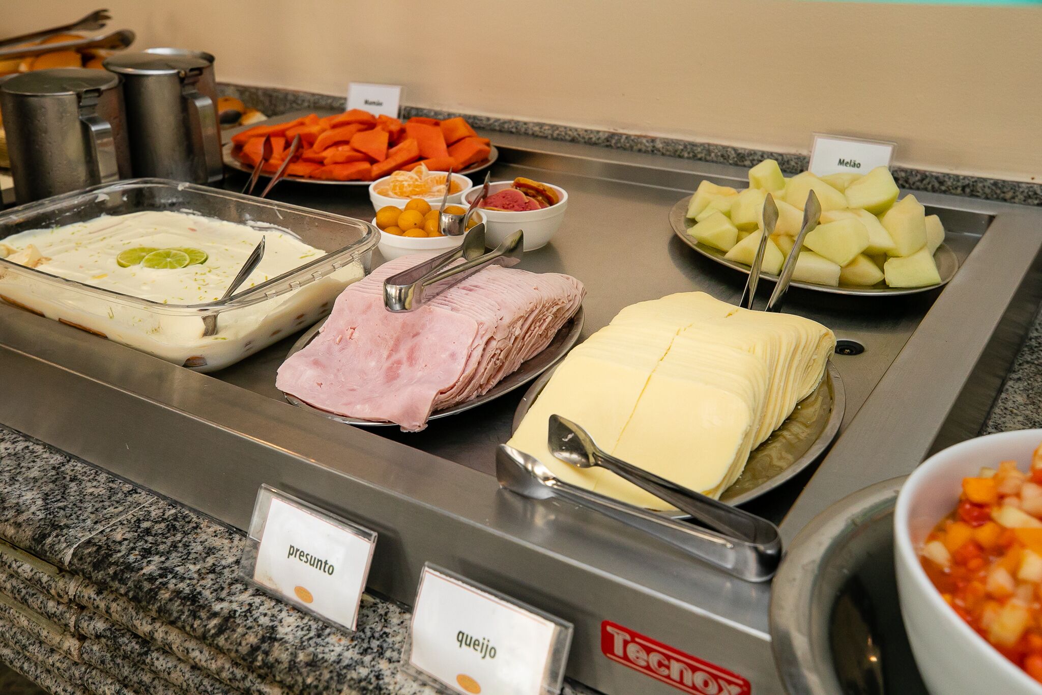 Un buffet con diversi piatti, tra cui uno con 'presunto', uno con 'queijo' e uno con 'melão'.