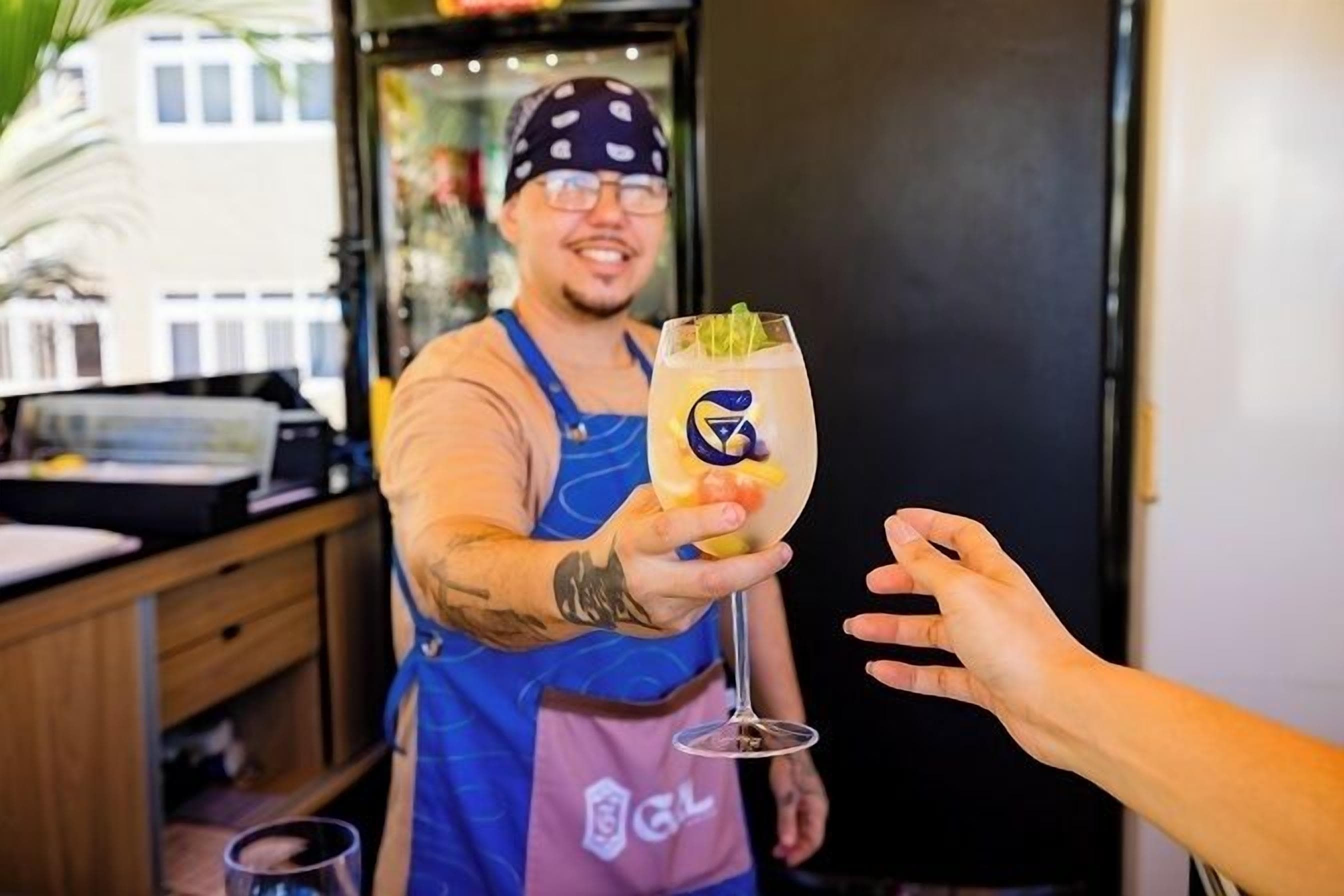 A person in a blue apron holding a drink with a logo, standing in a room with a counter and a door.