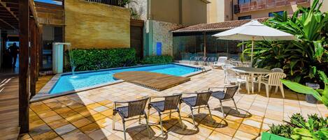 A swimming pool with lounge chairs and a table, surrounded by greenery and a tiled patio.