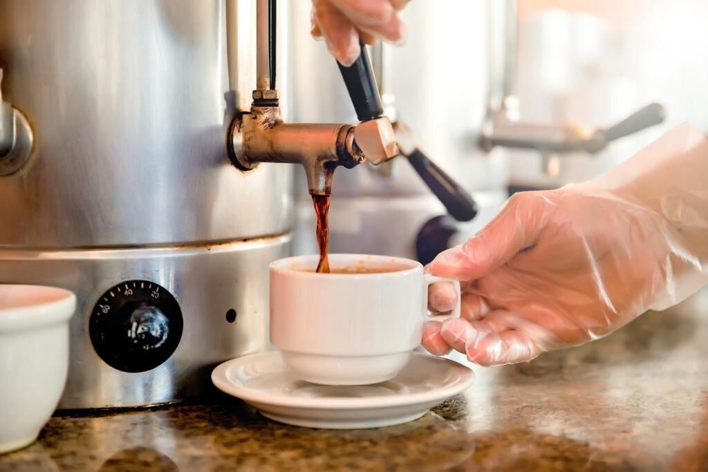 Eine Person in Handschuhen gießt Kaffee von einer Maschine in eine weiße Tasse mit Untertasse.