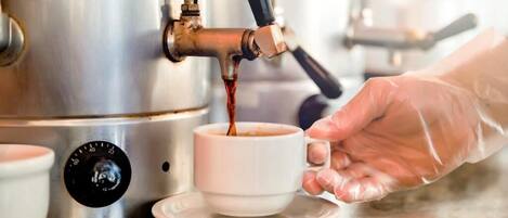 Eine Person in Handschuhen gießt Kaffee von einer Maschine in eine weiße Tasse mit Untertasse.