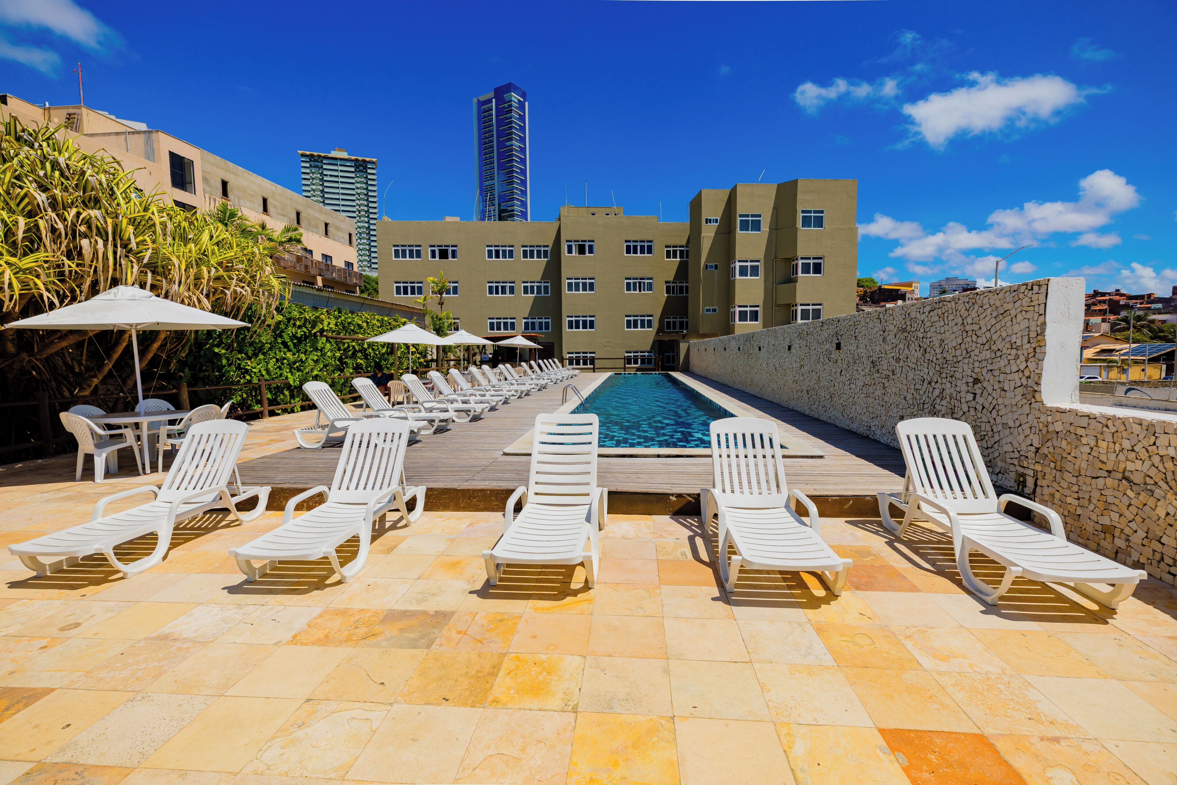 Zona junto a la piscina con sillones de descanso y sombrillas, una pared de piedra y una vista de un edificio alto al fondo.