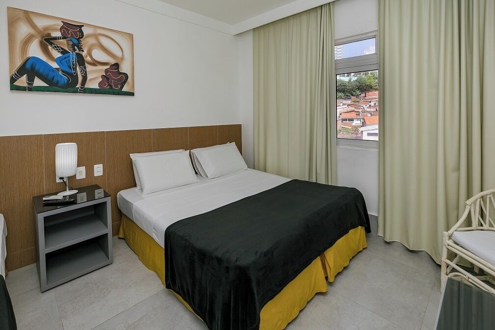 A hotel room with a bed, a nightstand, a chair, a window with curtains, and a painting on the wall.