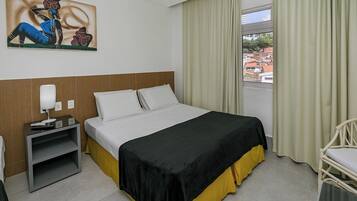 A hotel room with a bed, a nightstand, a chair, a window with curtains, and a painting on the wall.