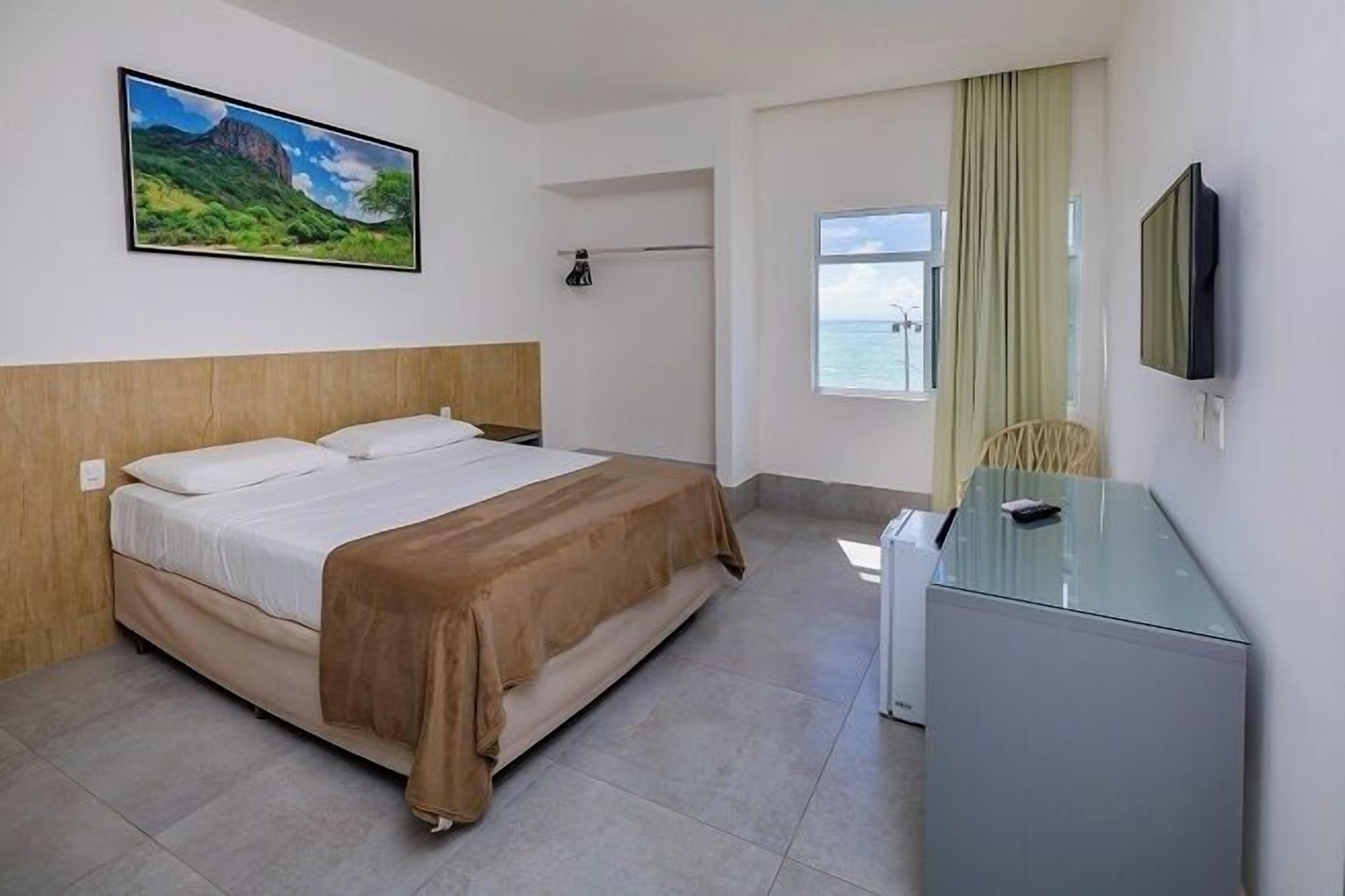 A hotel room with a bed, a desk, a TV, a window with a view, and a painting on the wall.