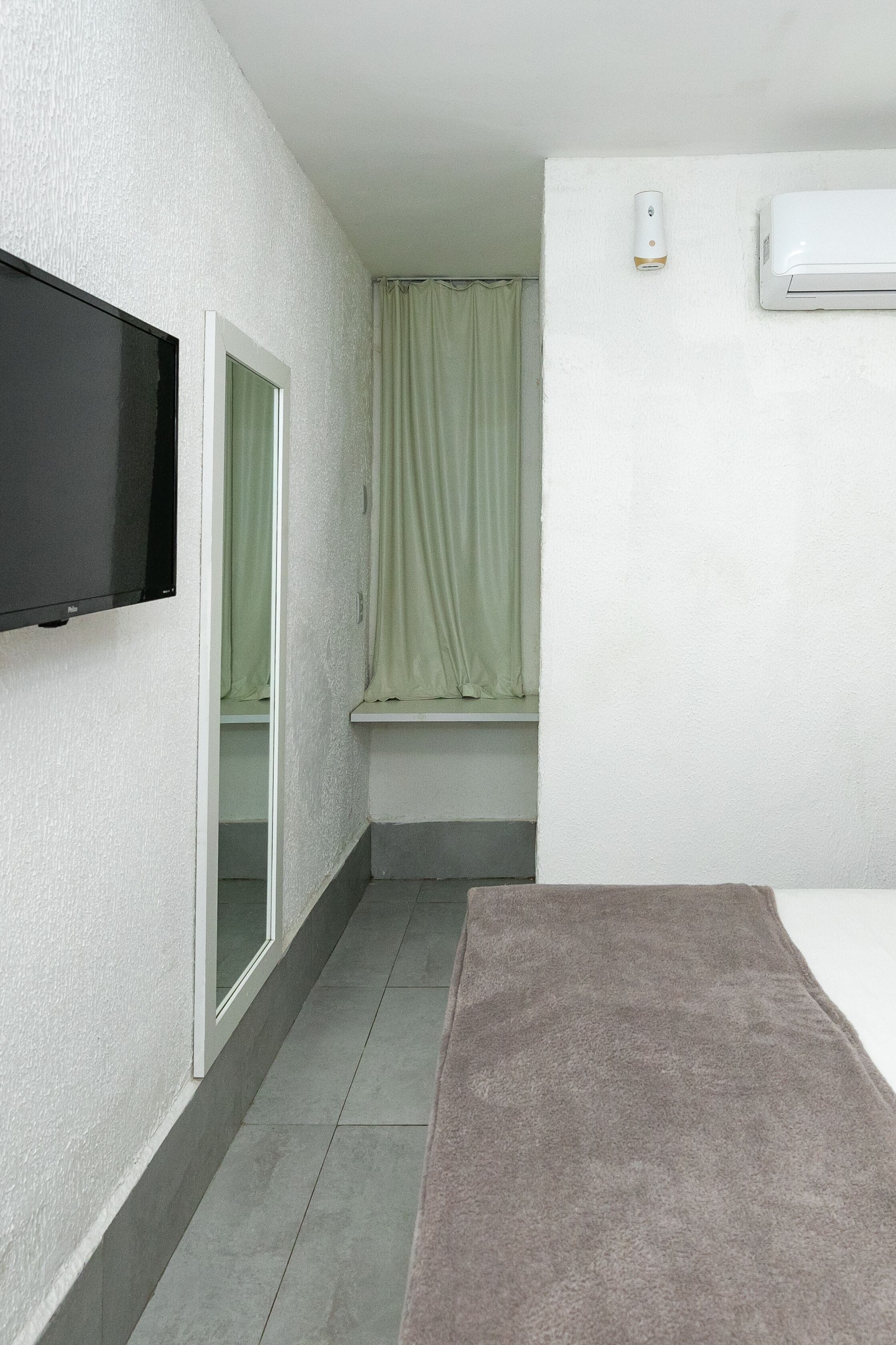 Una habitación pequeña y estrecha con una televisión de pantalla plana montada en la pared, una ventana con cortina verde y un aire acondicionado blanco en la pared.