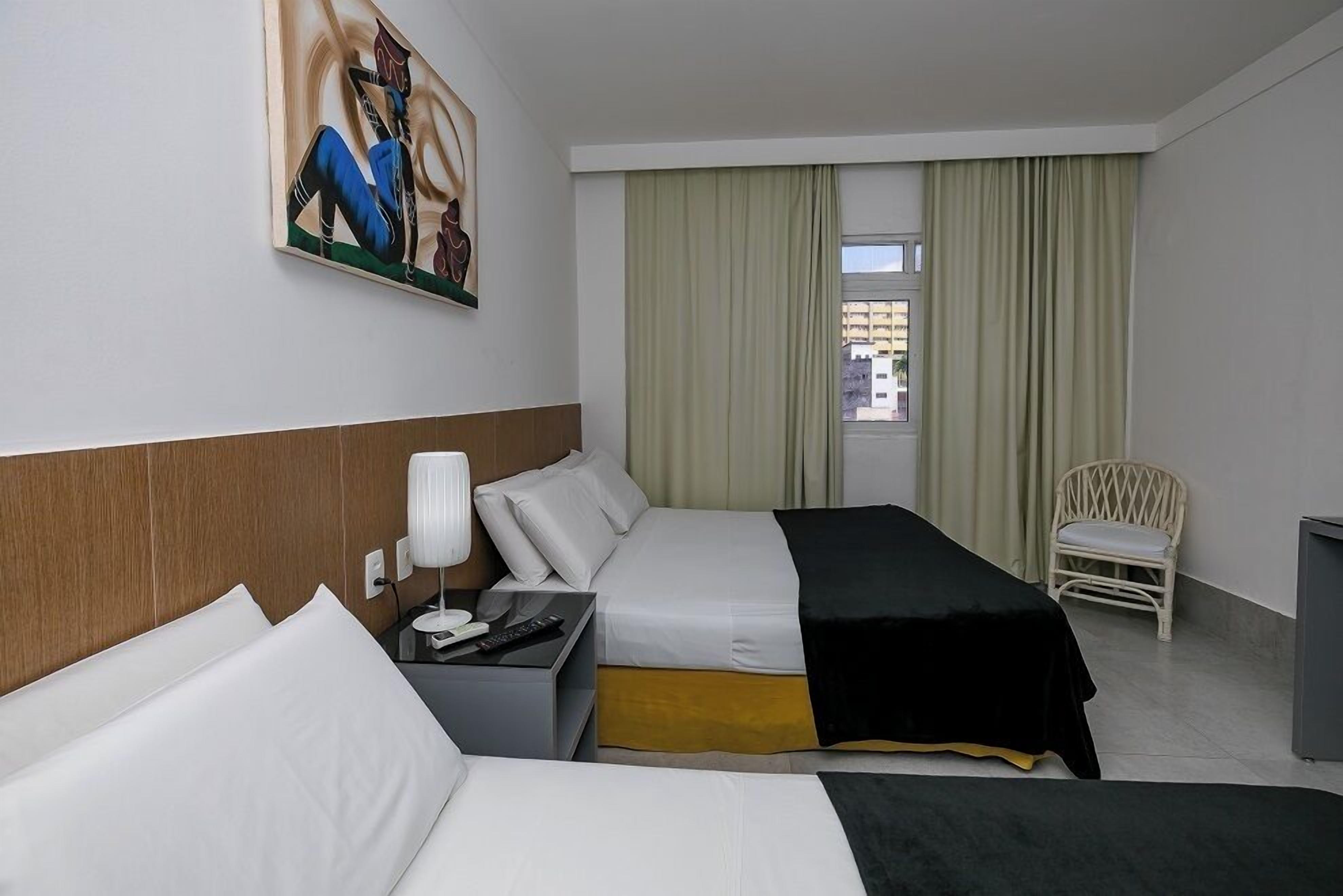 Una habitación de hotel con una cama, una silla, un cuadro y un ventanal con cortinas.