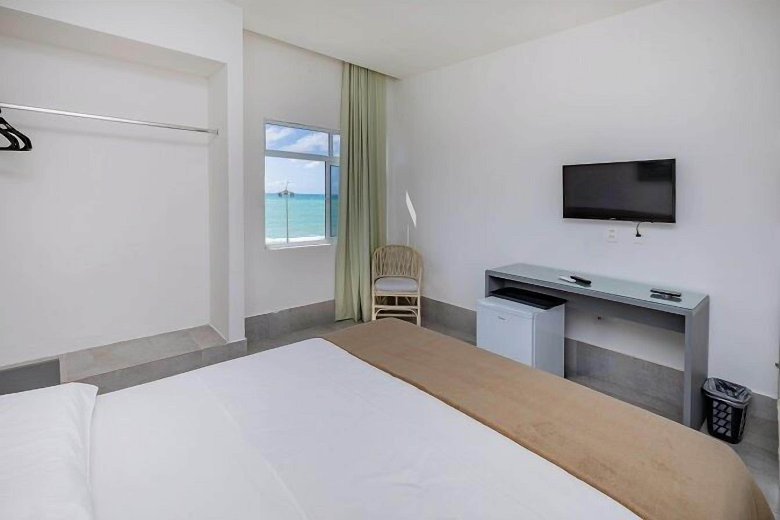 A hotel room with a bed, a desk, a chair, a TV, and a window with a view of the sea.