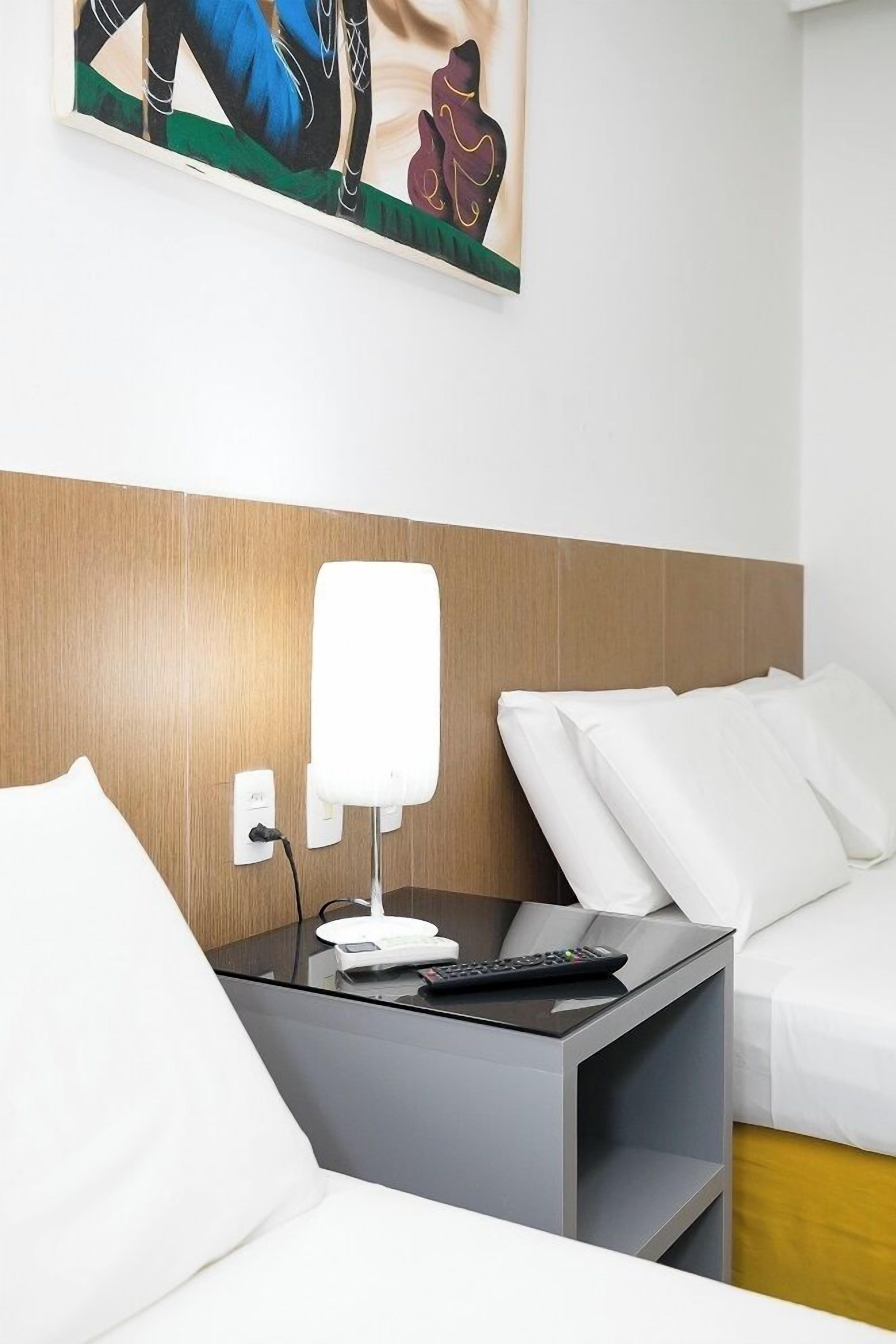 A hotel room with a bed, a bedside table with a lamp, a remote control, and a painting on the wall.