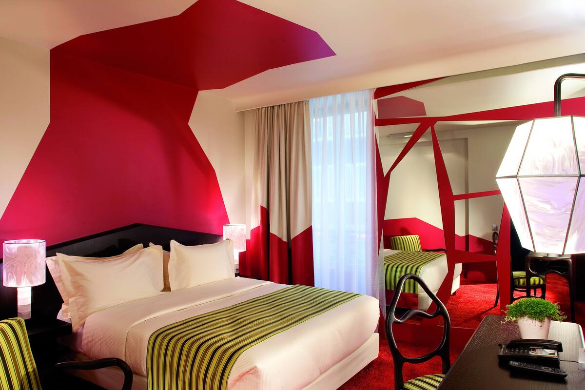 Une chambre d’hôtel avec un grand lit, un bureau avec une lampe, une chaise et une petite table sur laquelle est posée une plante.